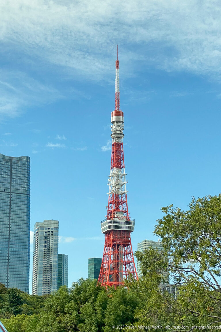 Tokyo Tower rises above trees and modern buildings beneath a bright blue sky.
