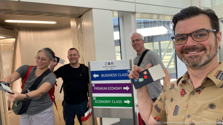 Four travelers pose beside business, premium economy, and economy class direction signs at an airport.