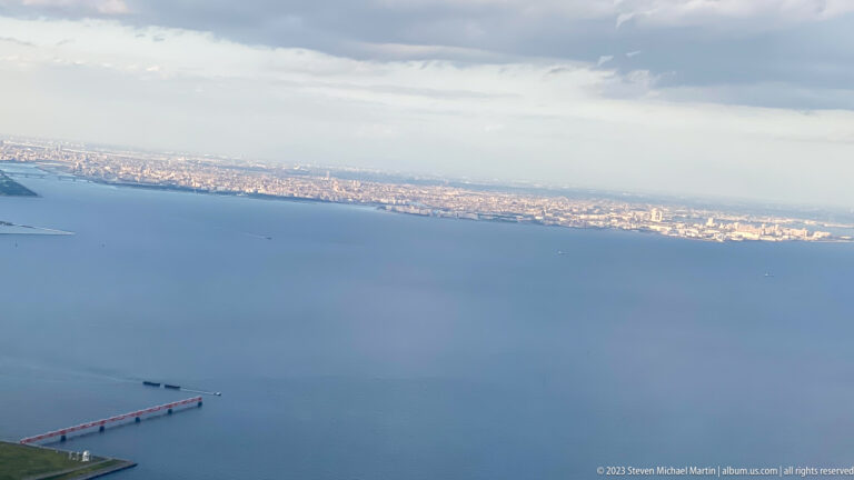 Aerial view over Tokyo Bay with a long narrow structure and distant city shoreline.