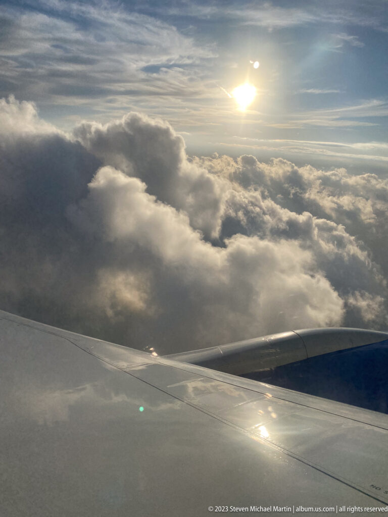 Aircraft wing beneath bright sun and billowing clouds viewed from above.