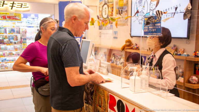 Two visitors speak with a masked attendant behind clear panels at a tourist information counter.