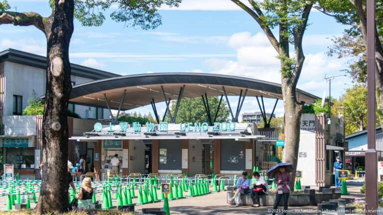 Curving entrance canopy at Ueno Zoo with bilingual lettering, visitors, and green barriers.