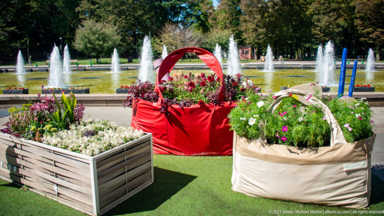 Red and white basket-shaped flower planters stand before a row of fountains in Ueno Park.