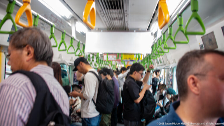 Passengers stand and sit inside a Tokyo train beneath green triangular hand straps.