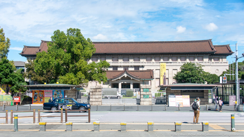 Tokyo National Museum entrance area with tiled roofs, gateposts, trees, and a larger building beyond.