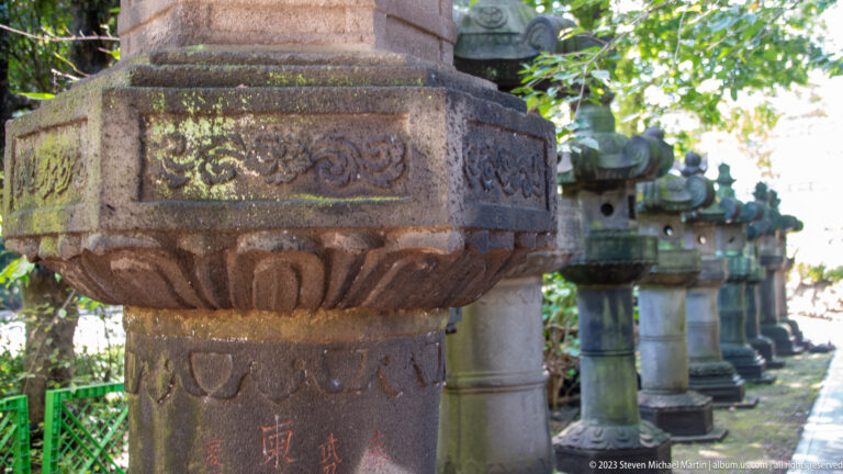 Close-up of a carved stone lantern base with lotus ornament, inscriptions, moss, and lanterns behind.