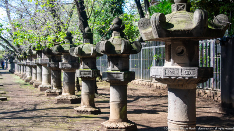 Long row of large weathered stone lanterns recedes through a shaded shrine precinct.