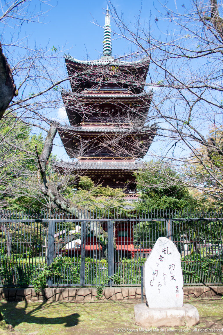Five-story wooden pagoda with tiled eaves and metal finial rises behind a fence and trees.