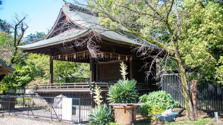 Small open-sided wooden pavilion with a broad roof stands among trees and fencing in Ueno Park.