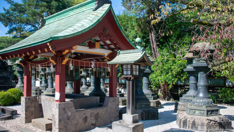 Roofed Shinto purification basin with hanging ladles, stone base, and lanterns in a garden setting.