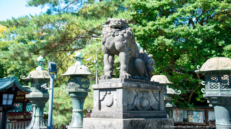 Stone komainu guardian on a pedestal surrounded by shrine lanterns and green trees.