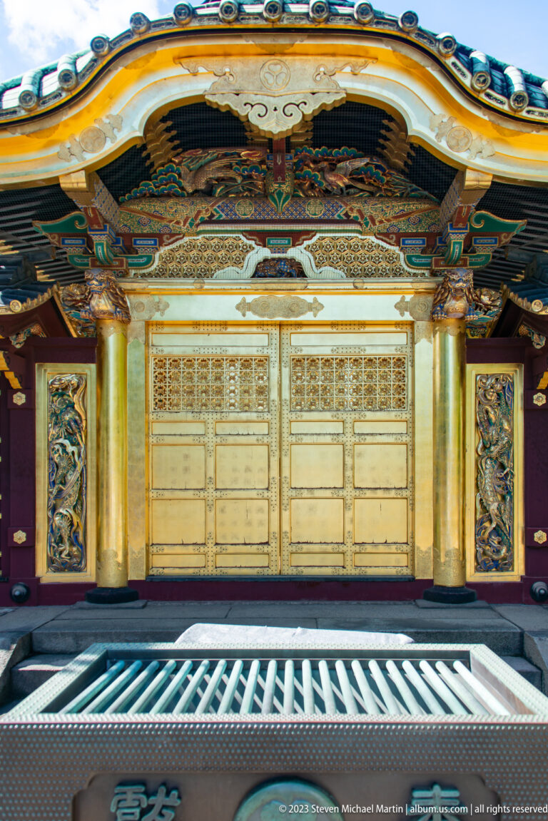 Frontal close view of gilded shrine doors, carved transom, painted reliefs, and decorated columns.