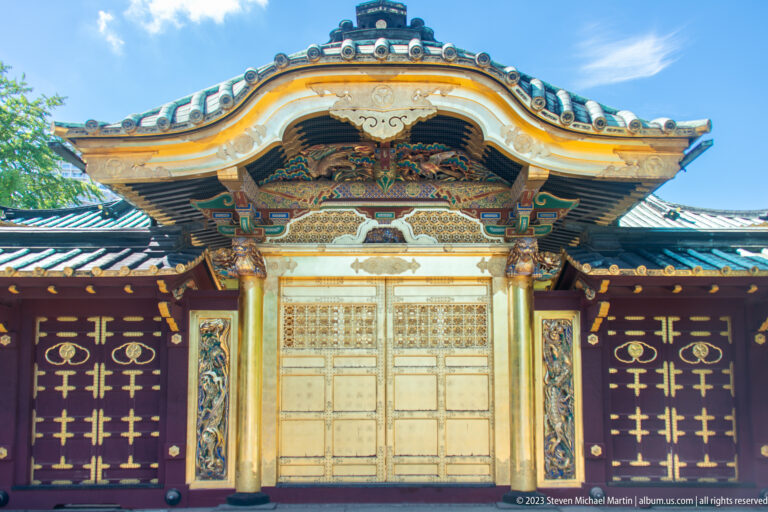 Full frontal view of an ornate shrine gate with gilded doors, curved roof, carvings, and dark side panels.