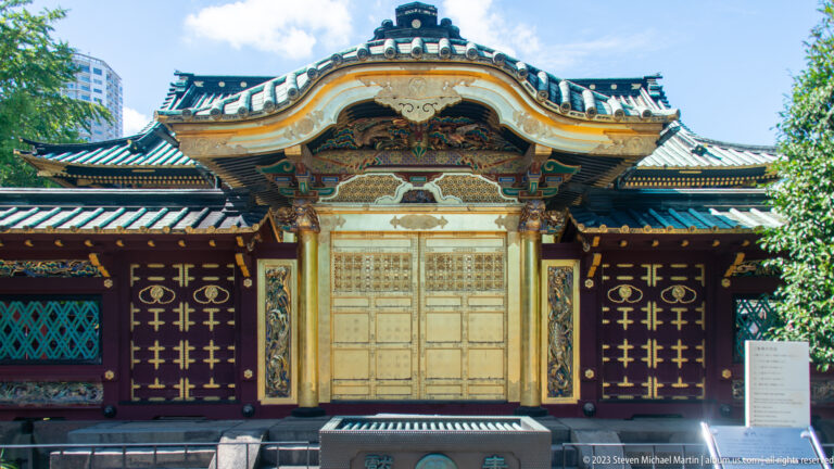 Wider frontal view of the gilded Karamon framed by shrine buildings, trees, and a visitor sign.