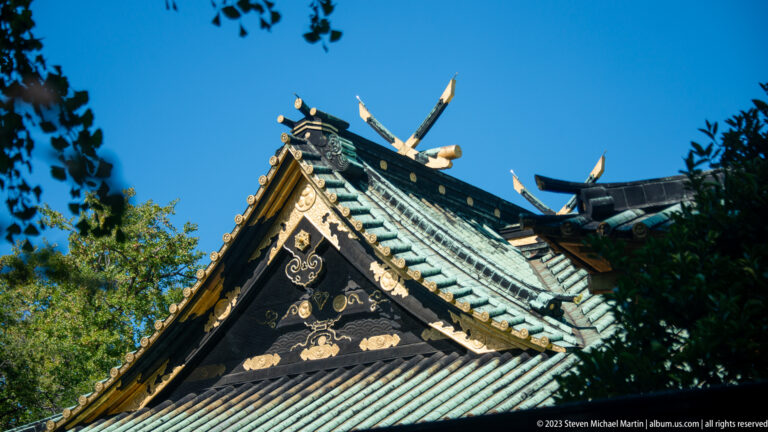 Upward view of dark shrine gables with green tiles, gold crests, and carved ornament against blue sky.