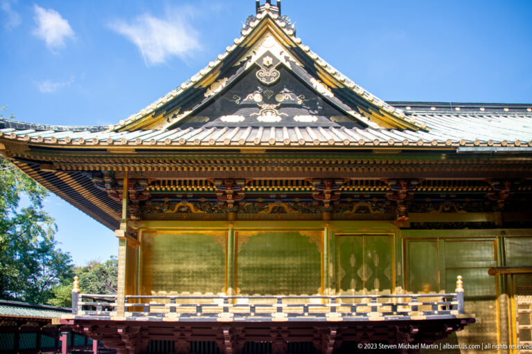 Side view of a shrine building with tiled gable, green shutters, gilded trim, and raised veranda.