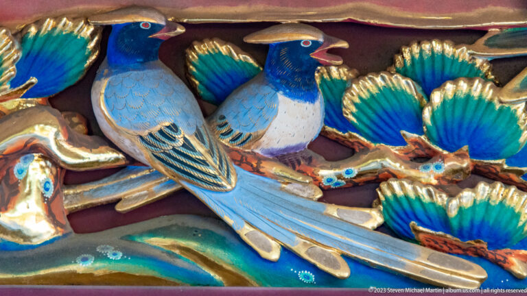 Close-up of a carved blue bird surrounded by green leaves, branches, and gold accents.