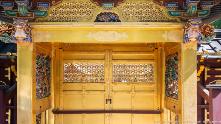 Inner view of a gilded shrine gate with heavy posts, lattice doors, carved brackets, and painted eaves.