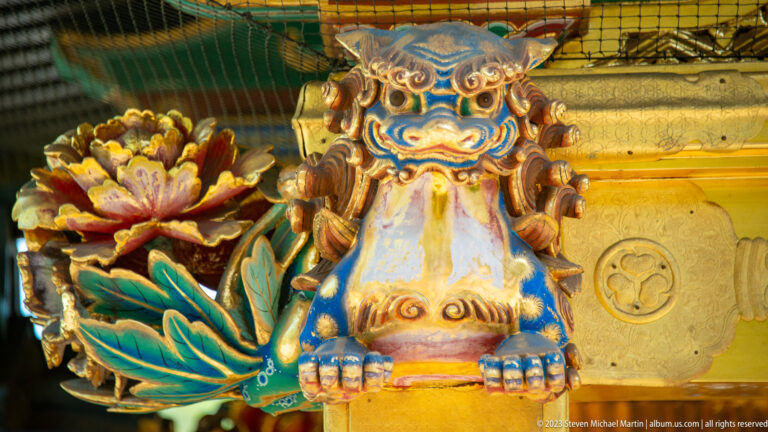 Colorful carved shishi with blue face beside a large peony blossom and green leaves.