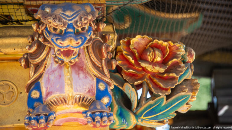 Blue-and-gold carved shishi with open mouth beside a red peony and green leaves.