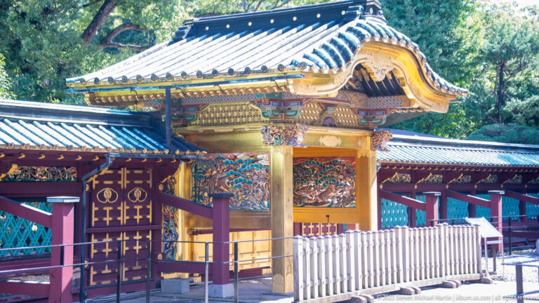 Angled view of an ornate shrine gate with curved roof, gilded posts, carved panels, and red walls.