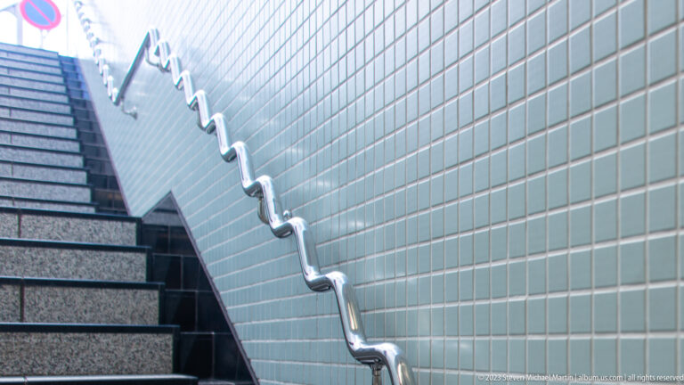 Polished metal handrail rises beside pale tiled wall and gray stairs in a modern stairwell.