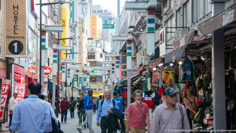 Pedestrians walk along a narrow Tokyo shopping lane lined with clothing stalls and signs.