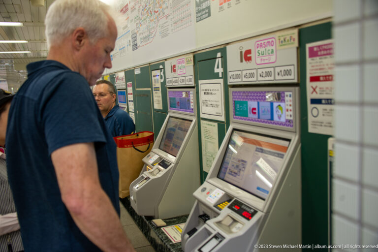 Passengers use green rail ticket machines beneath fare charts in a Tokyo station.
