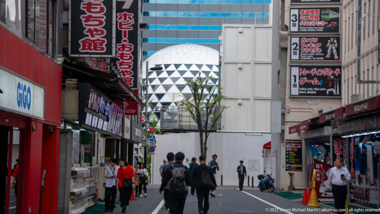 White diagrid Cocoon Tower appears between Shinjuku buildings above pedestrians and shop signs.