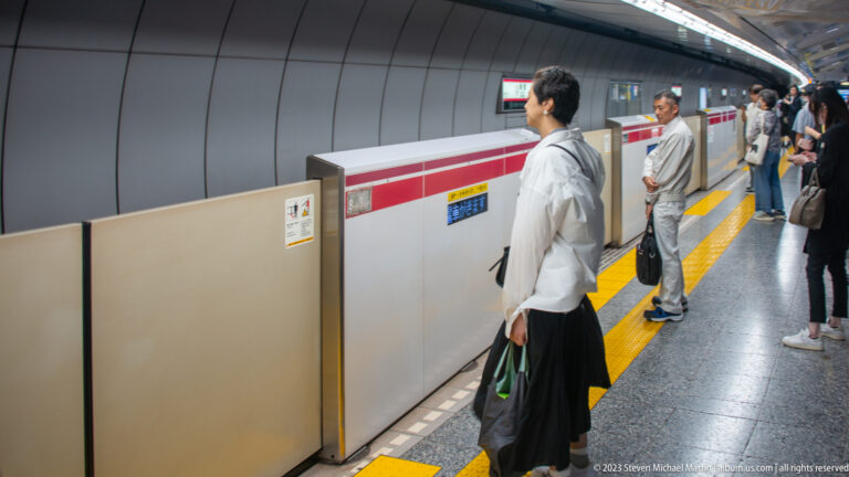 Passengers wait behind platform doors beside yellow tactile paving in a Tokyo subway station.