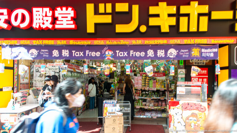Bright Don Quijote storefront with yellow lettering, tax-free signs, merchandise, and blurred pedestrians.