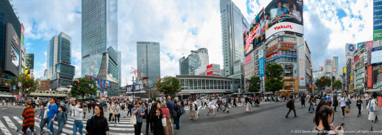Wide view of crowds crossing Shibuya beneath tall buildings, billboards, and a bright sky.