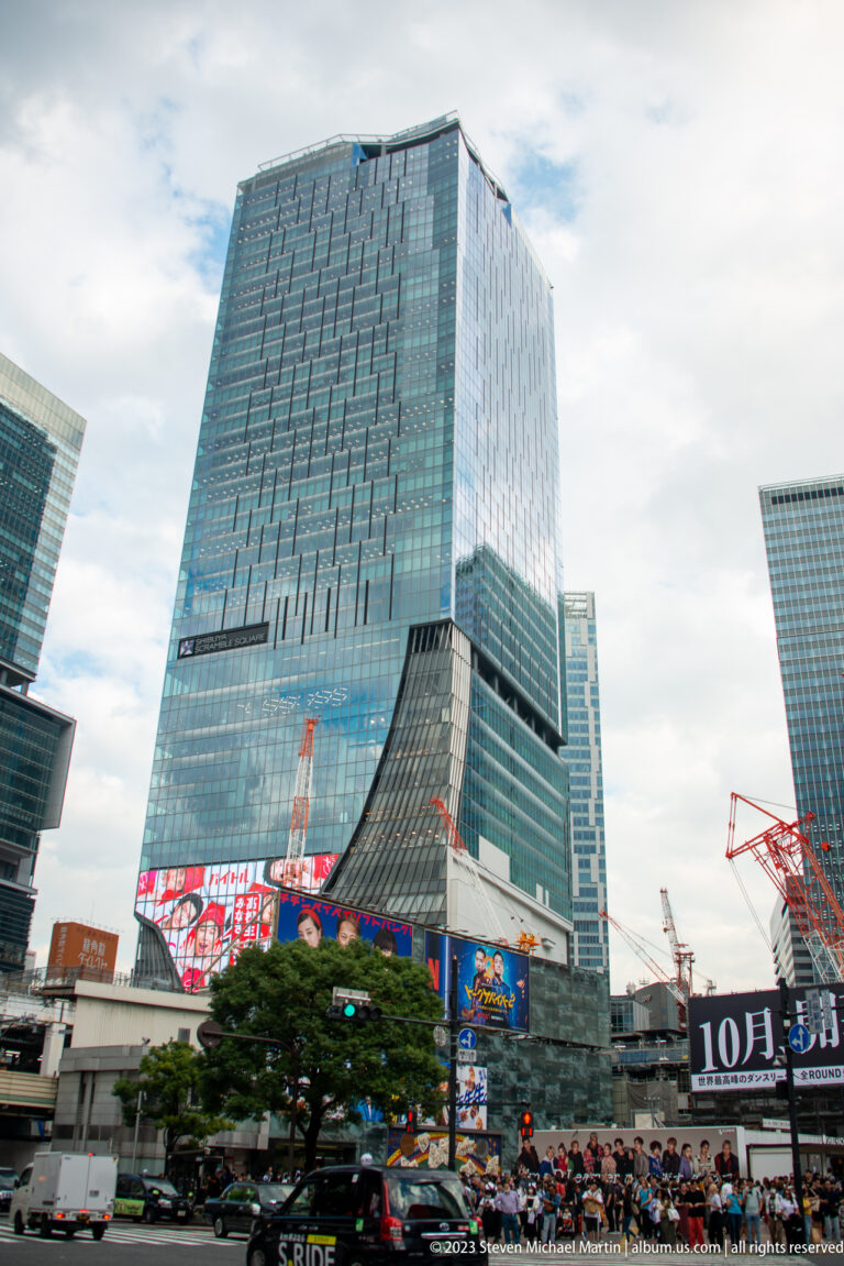 Glass Shibuya Scramble Square tower rises above station buildings, trees, signs, and cranes.