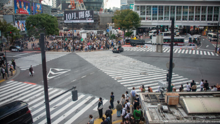 Elevated view of Shibuya Crossing with cars in the intersection and crowds waiting at the curb.