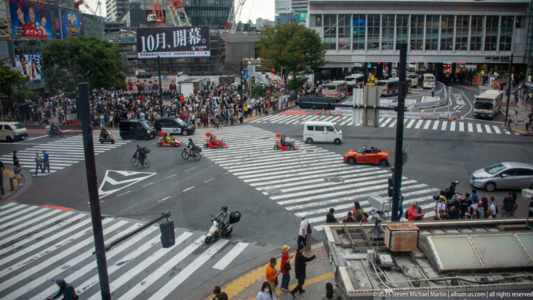 Cars and buses move through Shibuya Crossing while pedestrians gather along the sidewalks.