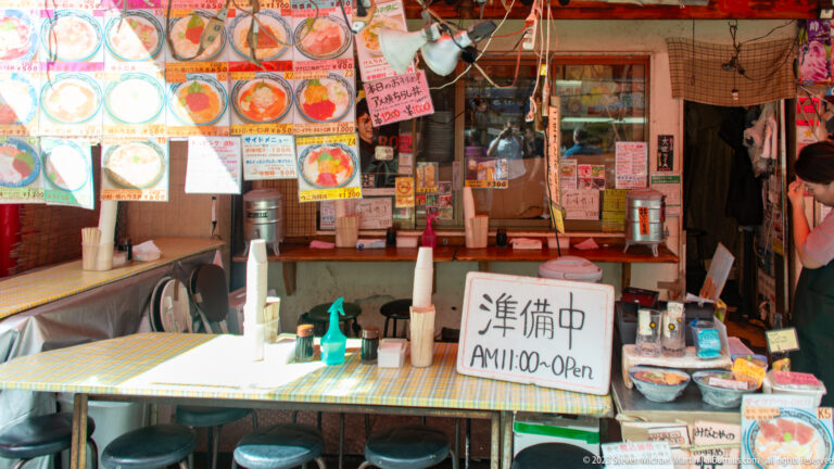 Small open-front restaurant with counter stools, menu photographs, bowls, and a preparing sign.