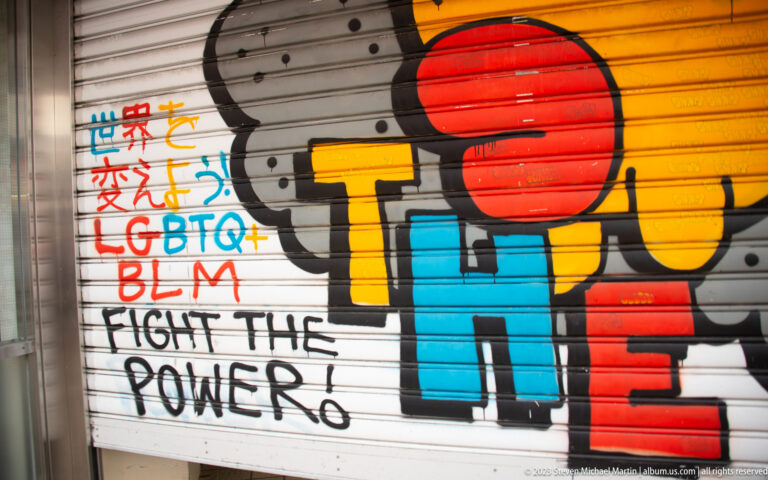 Colorful mural on a metal shutter with red sun, block letters, LGBTQ+, BLM, and Fight the Power text.