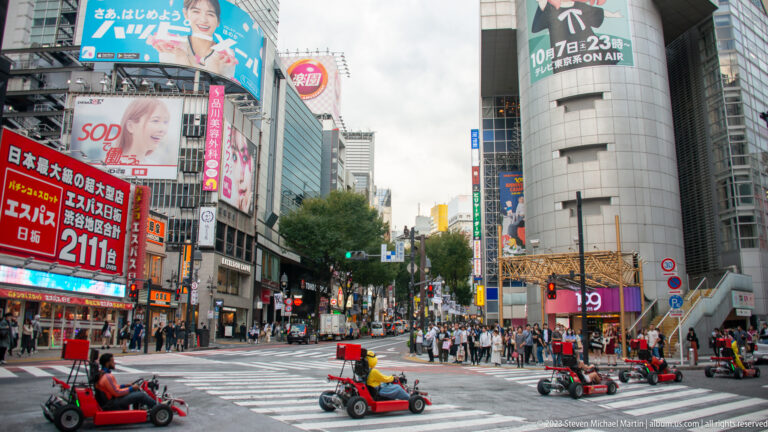 Several people drive small red mobility scooters across a wide Shibuya intersection.