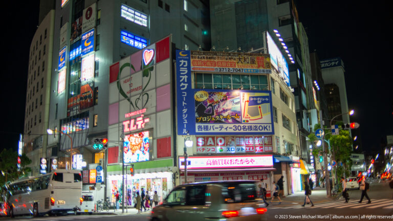 Night intersection with bright karaoke signs, lit storefronts, cars, and pedestrians.