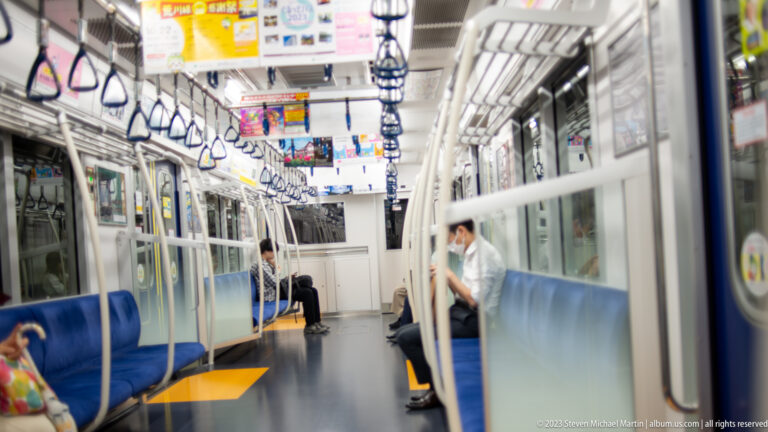 Nearly empty train carriage with blue bench seats, hanging straps, glass partitions, and seated passengers.