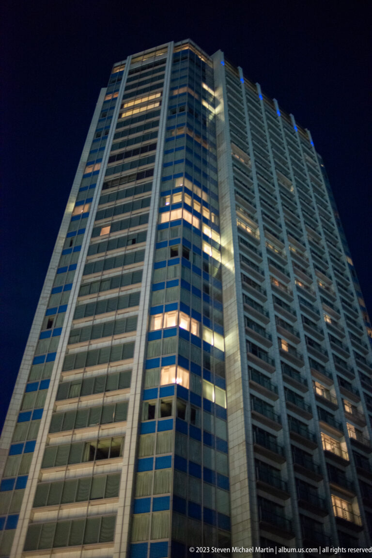 Low-angle night view of a tall modern tower with repeated windows and several illuminated rooms.