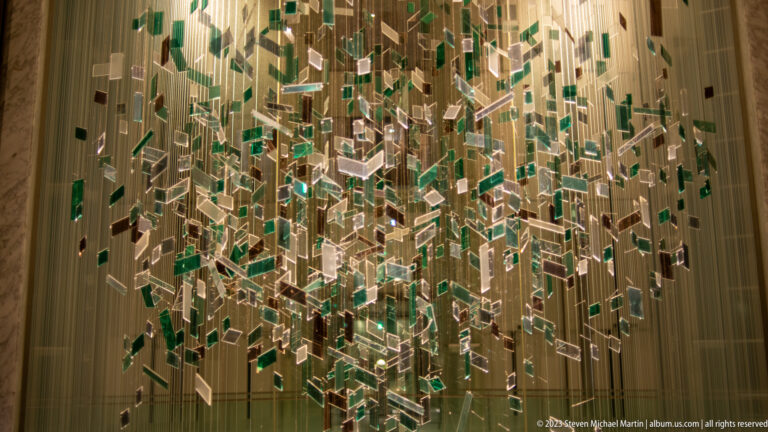 Suspended installation of many clear and green glass rectangles glowing under warm interior light.