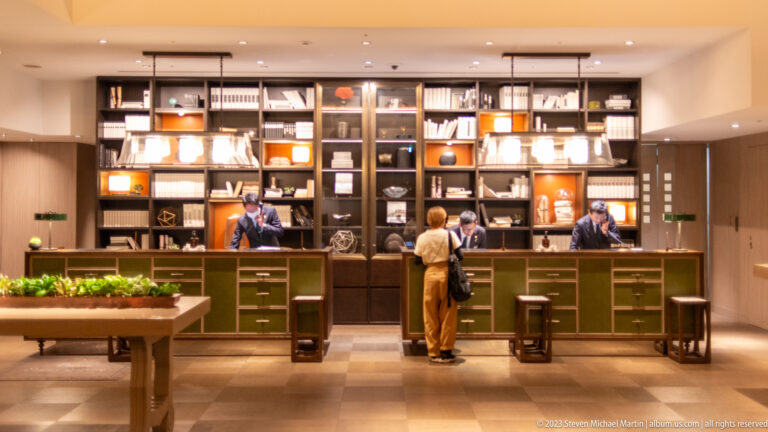 Guest speaks with staff at a green hotel reception desk backed by illuminated shelves and books.