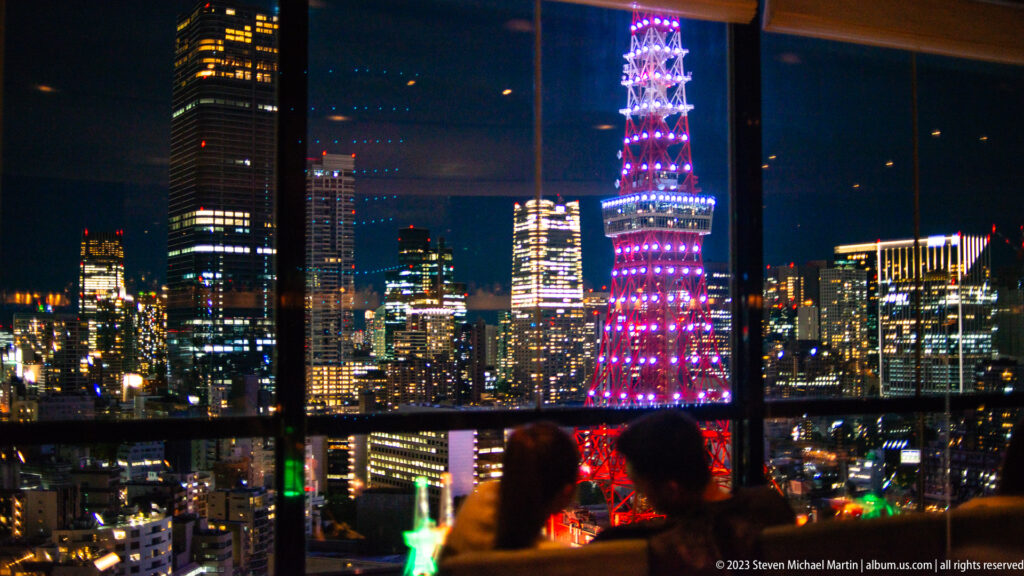 Illuminated Tokyo Tower seen through a high-rise window with two silhouetted viewers in foreground.