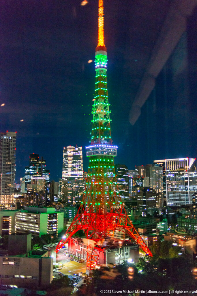 Tokyo Tower glows green, red, and orange above surrounding city buildings at night.