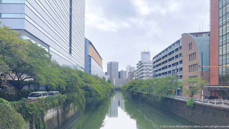 Calm narrow canal lined with trees, retaining walls, and modern buildings under cloudy sky.