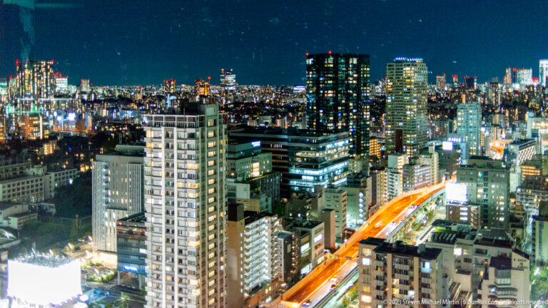 Elevated Tokyo night view with dense buildings and a curving orange-lit road.
