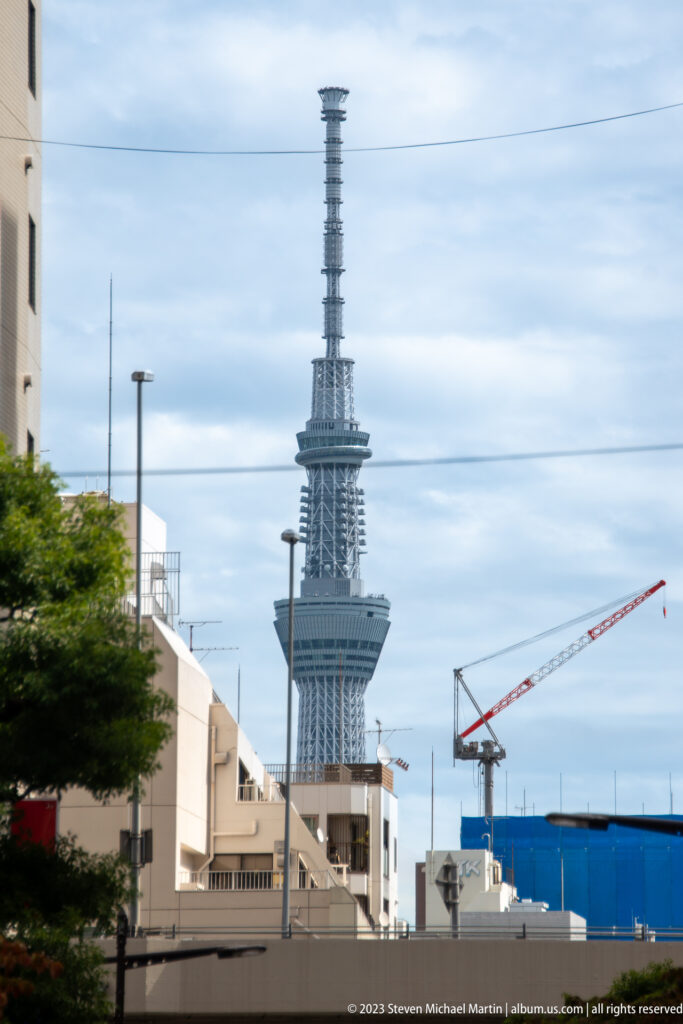 Tokyo Skytree rises beyond low buildings, utility lines, trees, and a construction crane.
