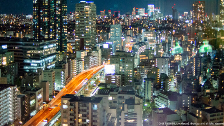 Elevated Tokyo night skyline with dense buildings and an orange-lit expressway.