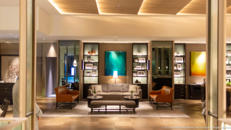 Modern lounge with gray sofa, brown armchairs, shelves, and colorful artwork.
